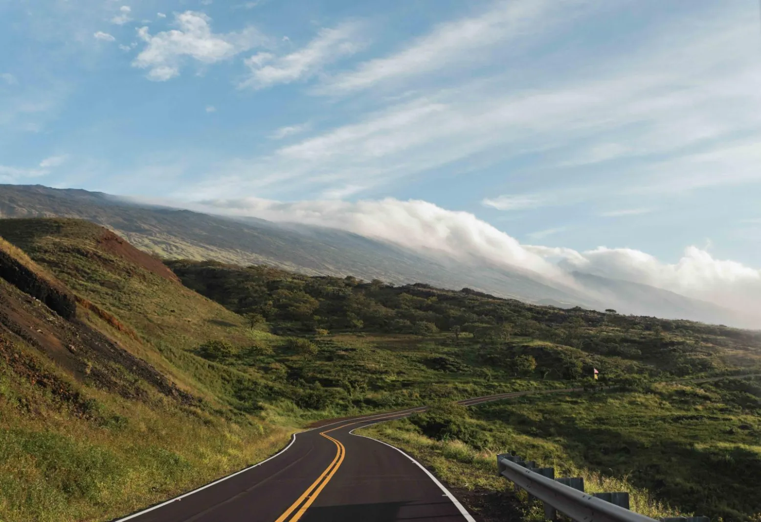A two-lane road curving across the open upland flank of Haleakala with cloud spilling over the ridge
