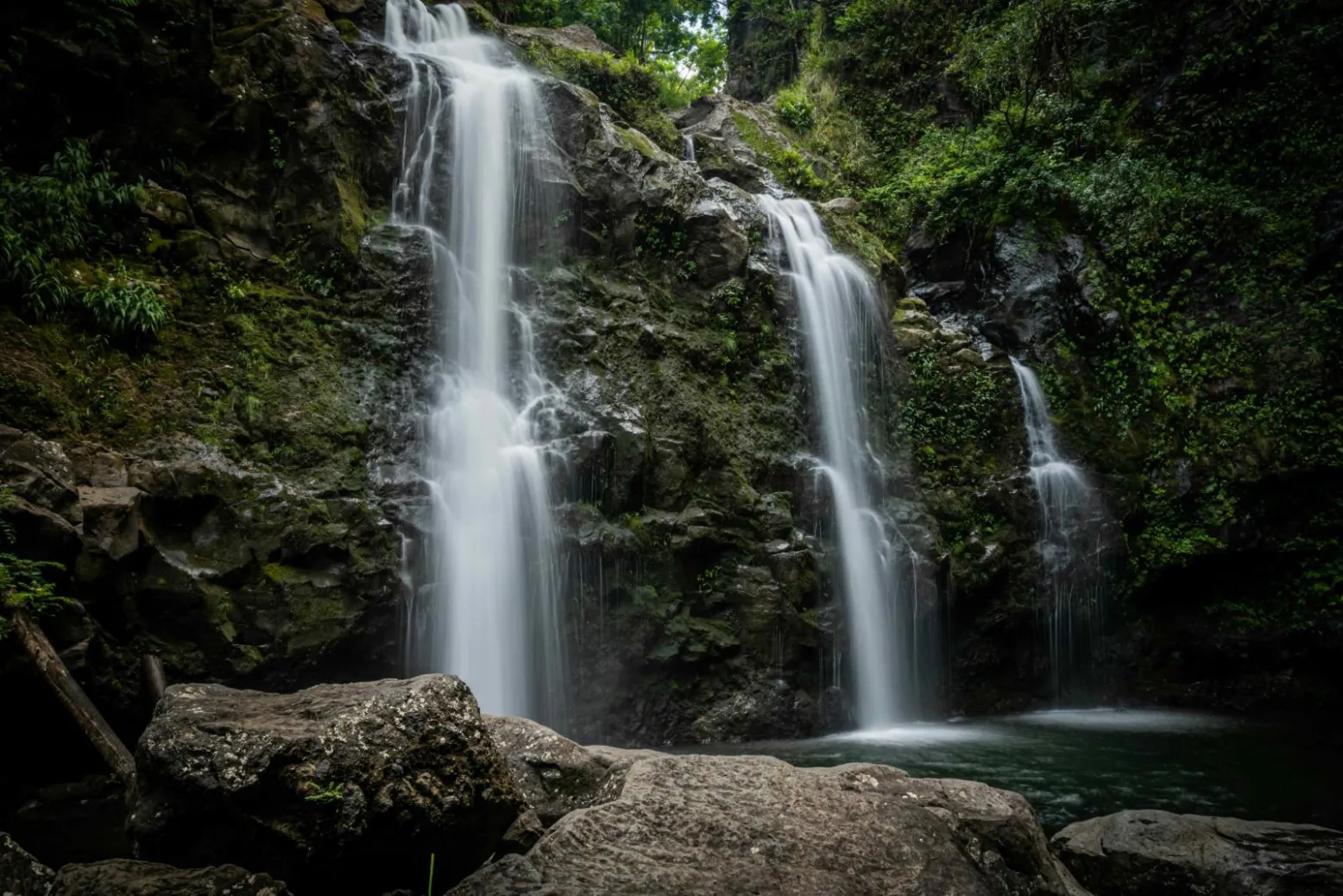 Three waterfalls dropping side by side into a pool surrounded by dark rock and green growth