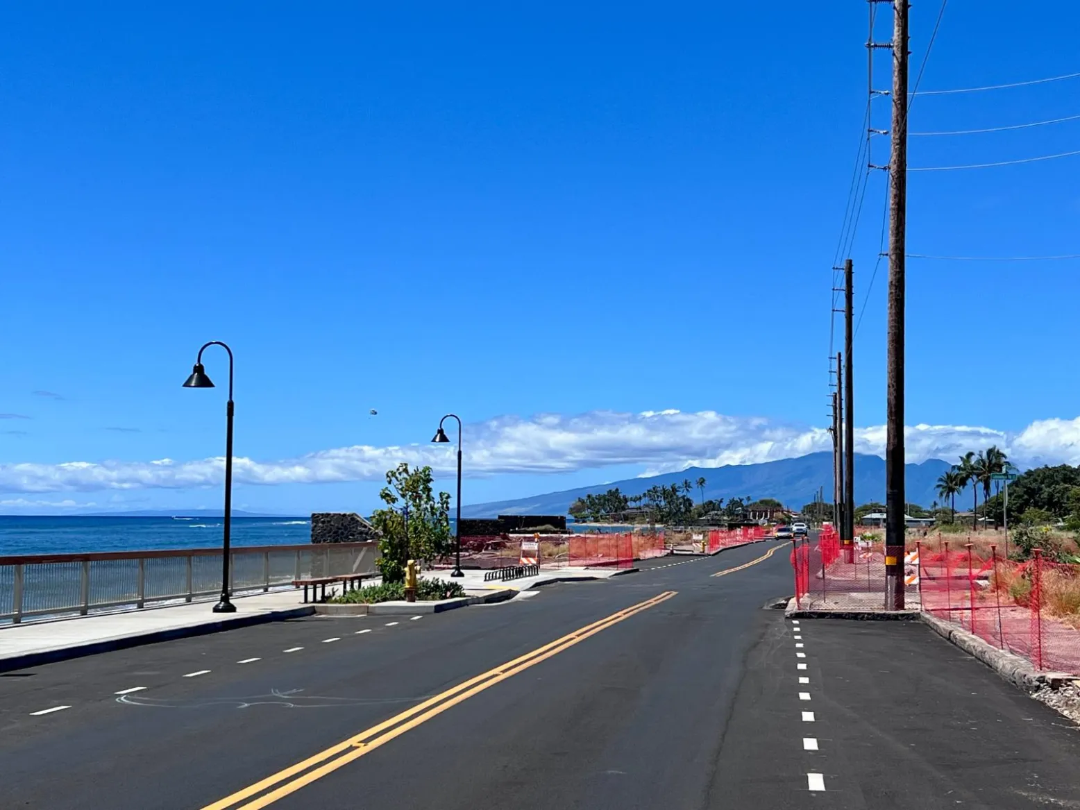 A repaved Front Street in Lāhainā beside the seawall, with new lamp posts, orange construction fencing and cleared lots