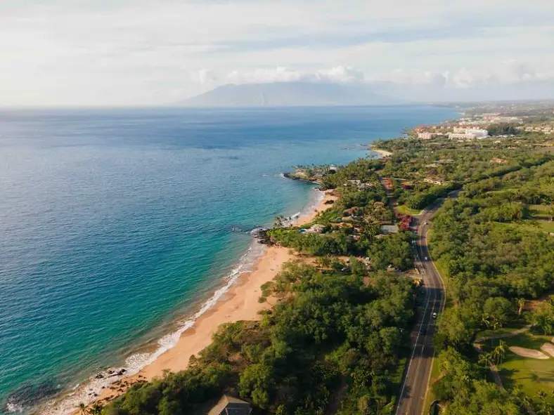 Aerial view of a South Maui road running between green hillside and a sandy cove
