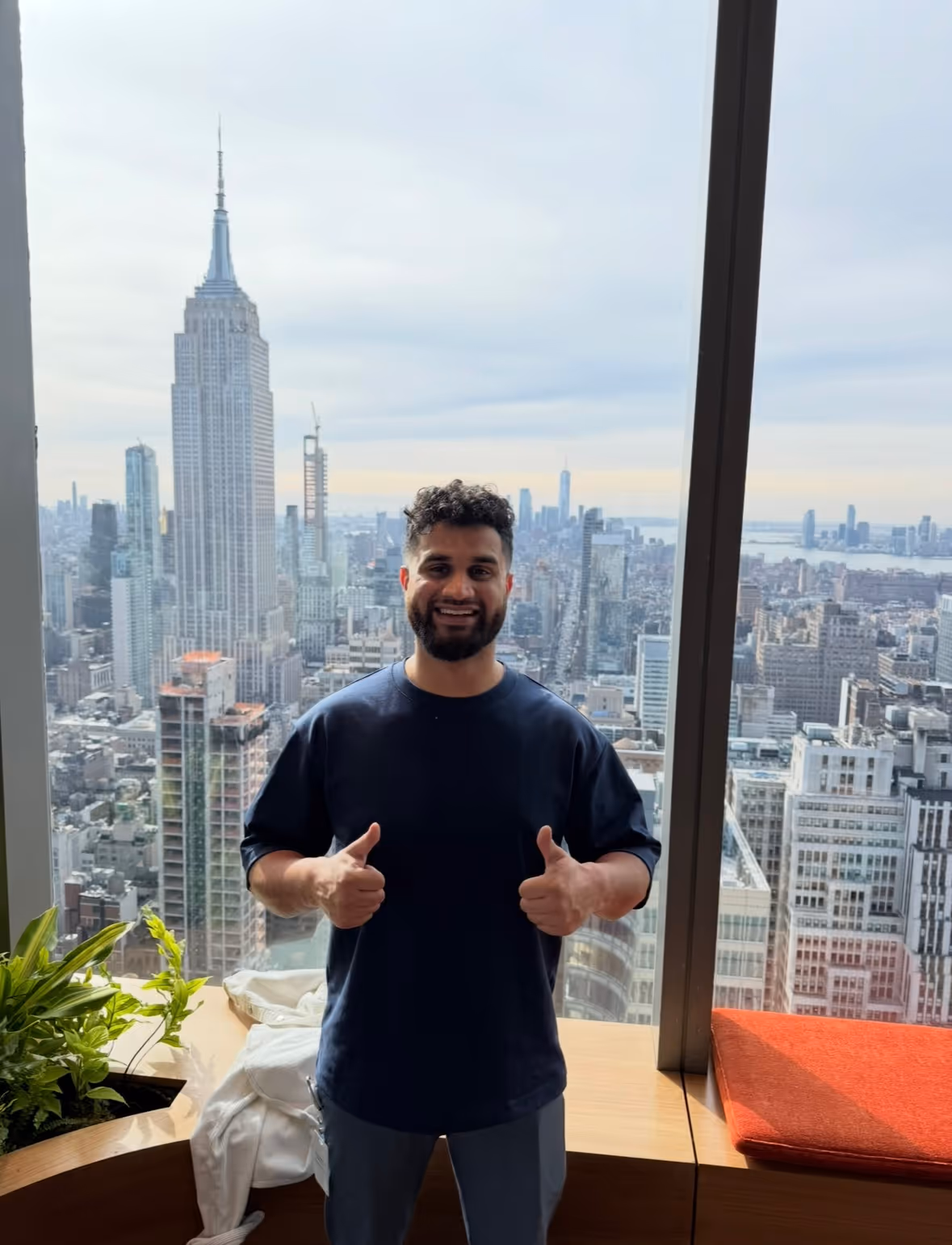 Smiling man with beard showing thumbs up in front of a large window with a city skyline including the Empire State Building.