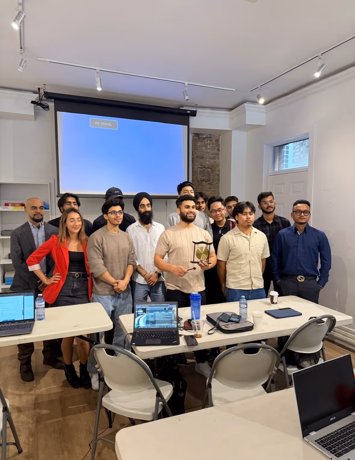 Group of people posing indoors with laptops on tables and a man holding a small gong in front of a projector screen.