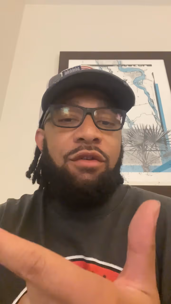 Man with glasses, a beard, and a cap featuring an American flag patch gesturing with his hand.