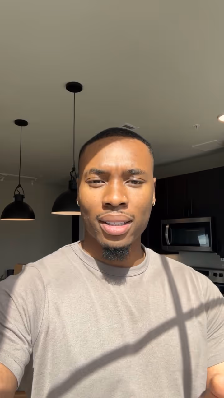 Young man with a short beard and earrings wearing a beige shirt in a sunlit modern kitchen.