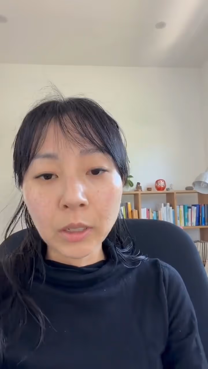 Woman with black hair and a black shirt sitting in front of a bookshelf with books and decorative items.