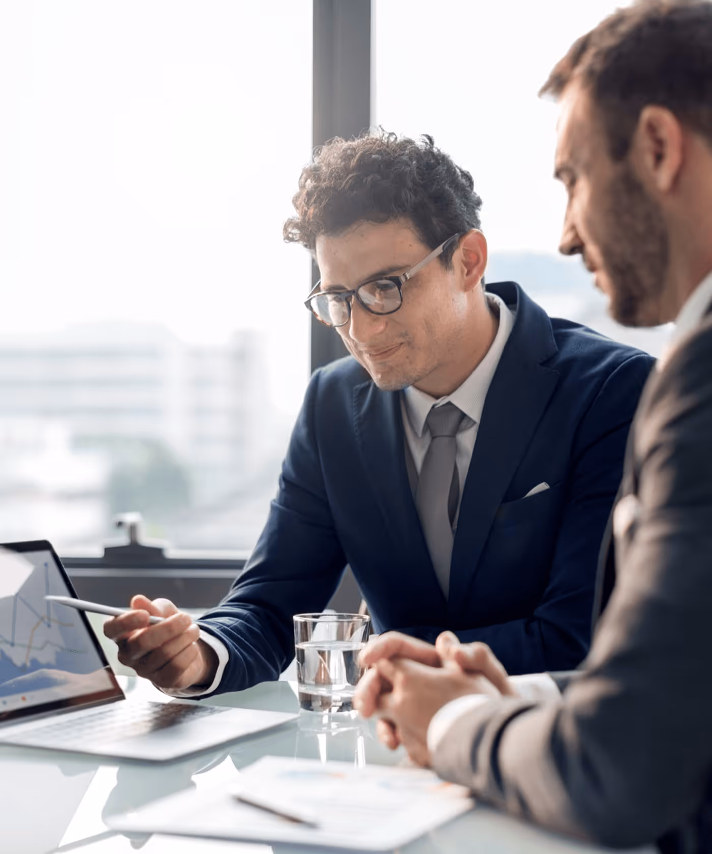 Geschäftsmänner diskutieren über Laptop-Statistiken in hellem Büro, einer deutet mit Stift auf den Bildschirm.