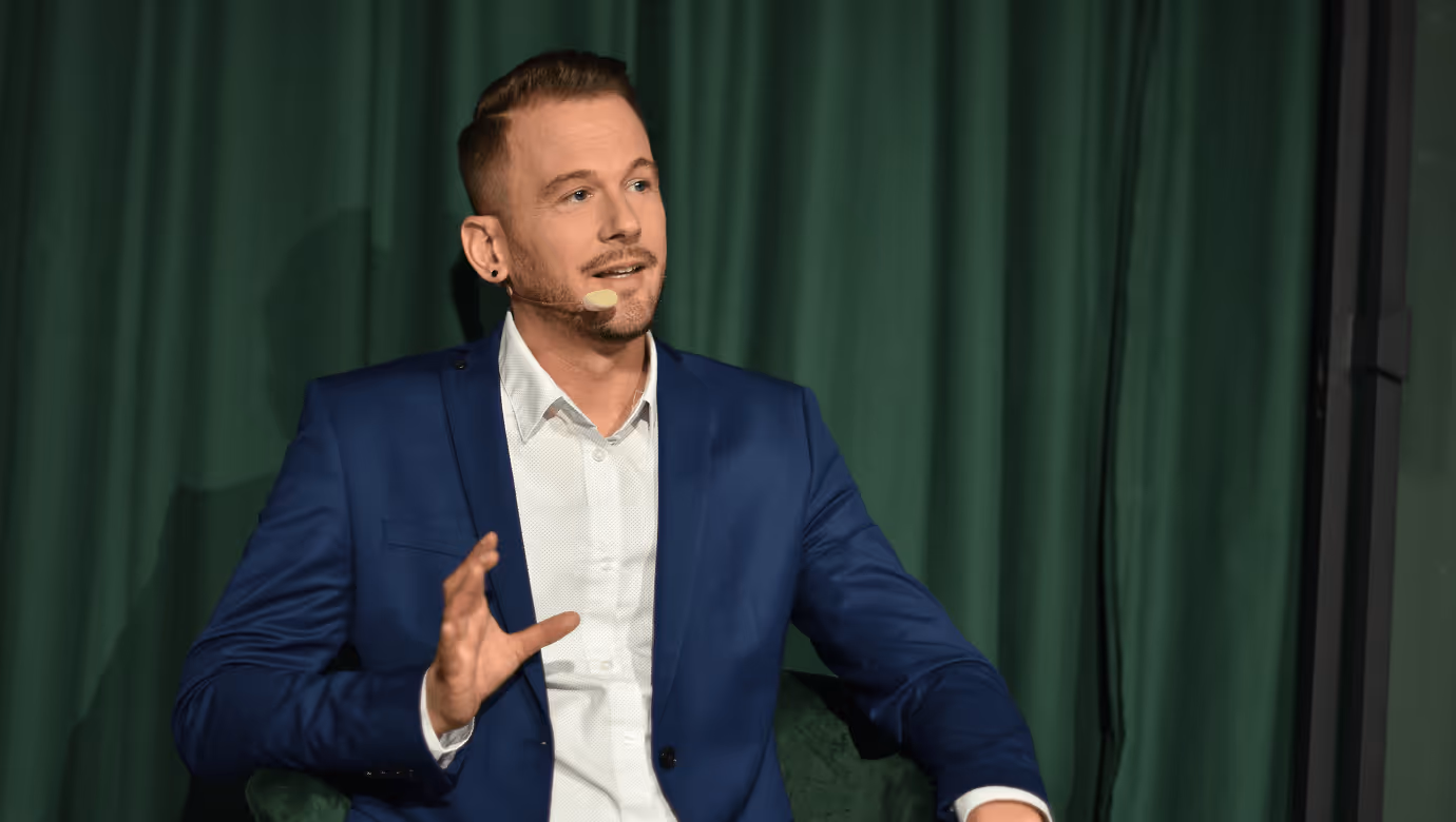 Mann in blauem Anzug diskutiert gestikulierend vor grünem Vorhang.