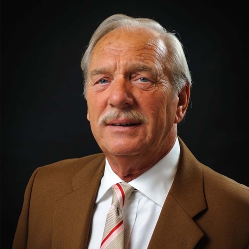 Smiling man with dark hair wearing a gray button-up shirt against a plain gray background.