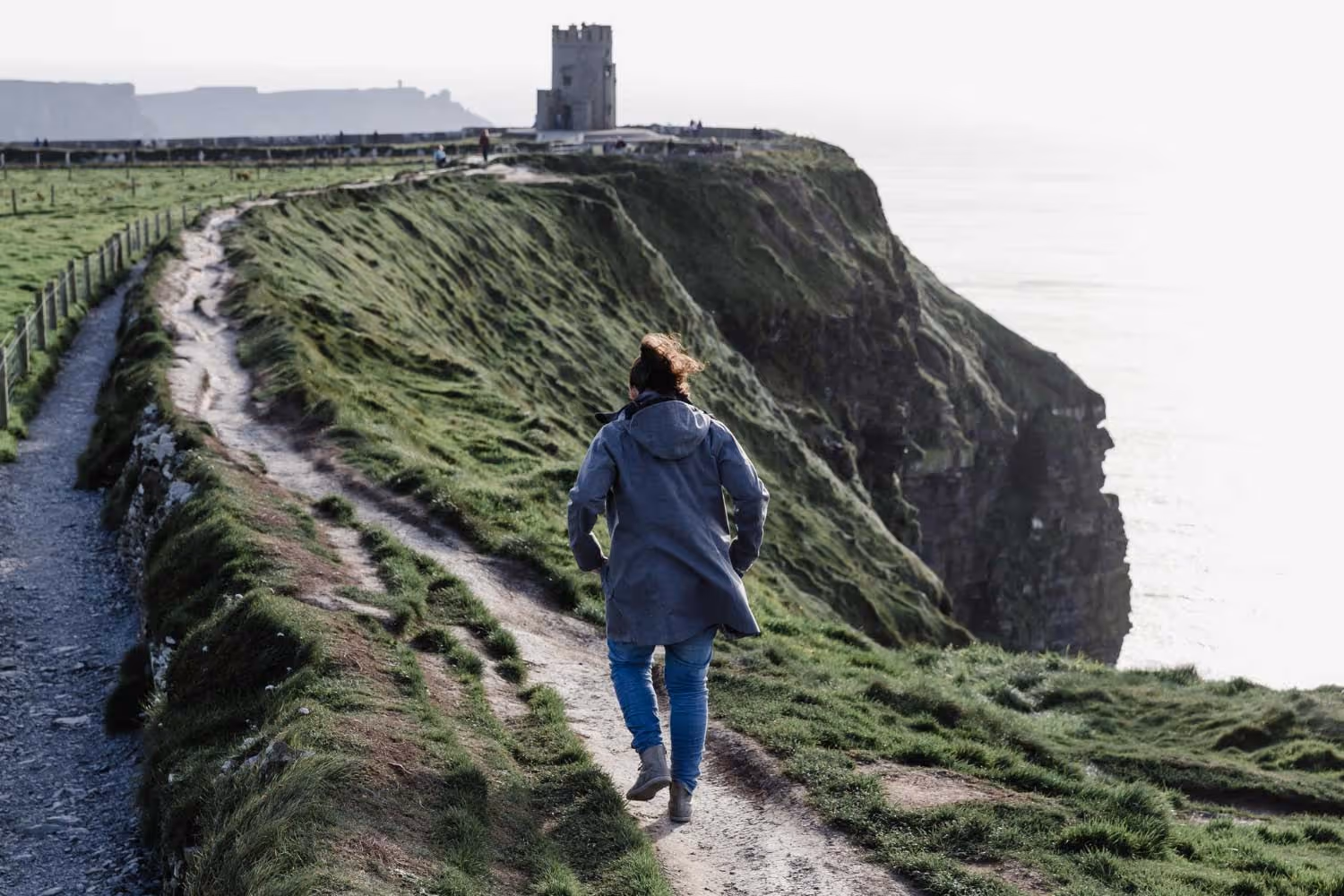 Person walking on a grassy cliffside path near a stone tower overlooking the ocean.