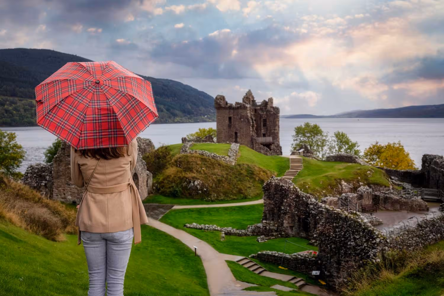 Person with red plaid umbrella overlooking Urquhart Castle ruins by a lake and hills.