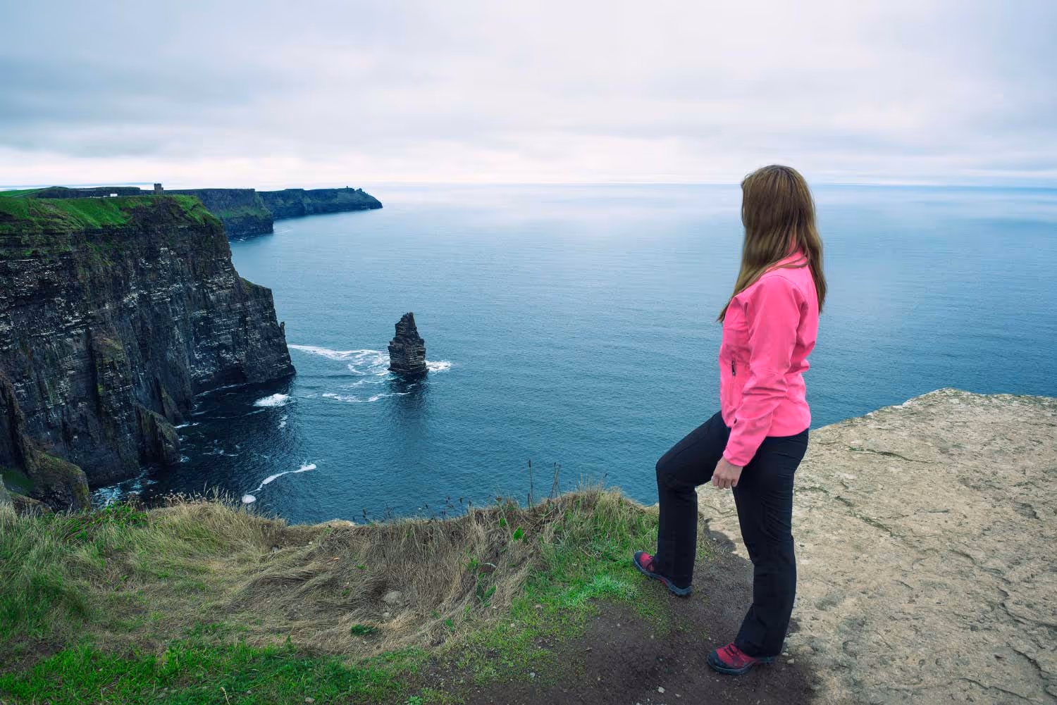 Woman in pink jacket standing on cliff edge overlooking ocean and rocky cliffs.