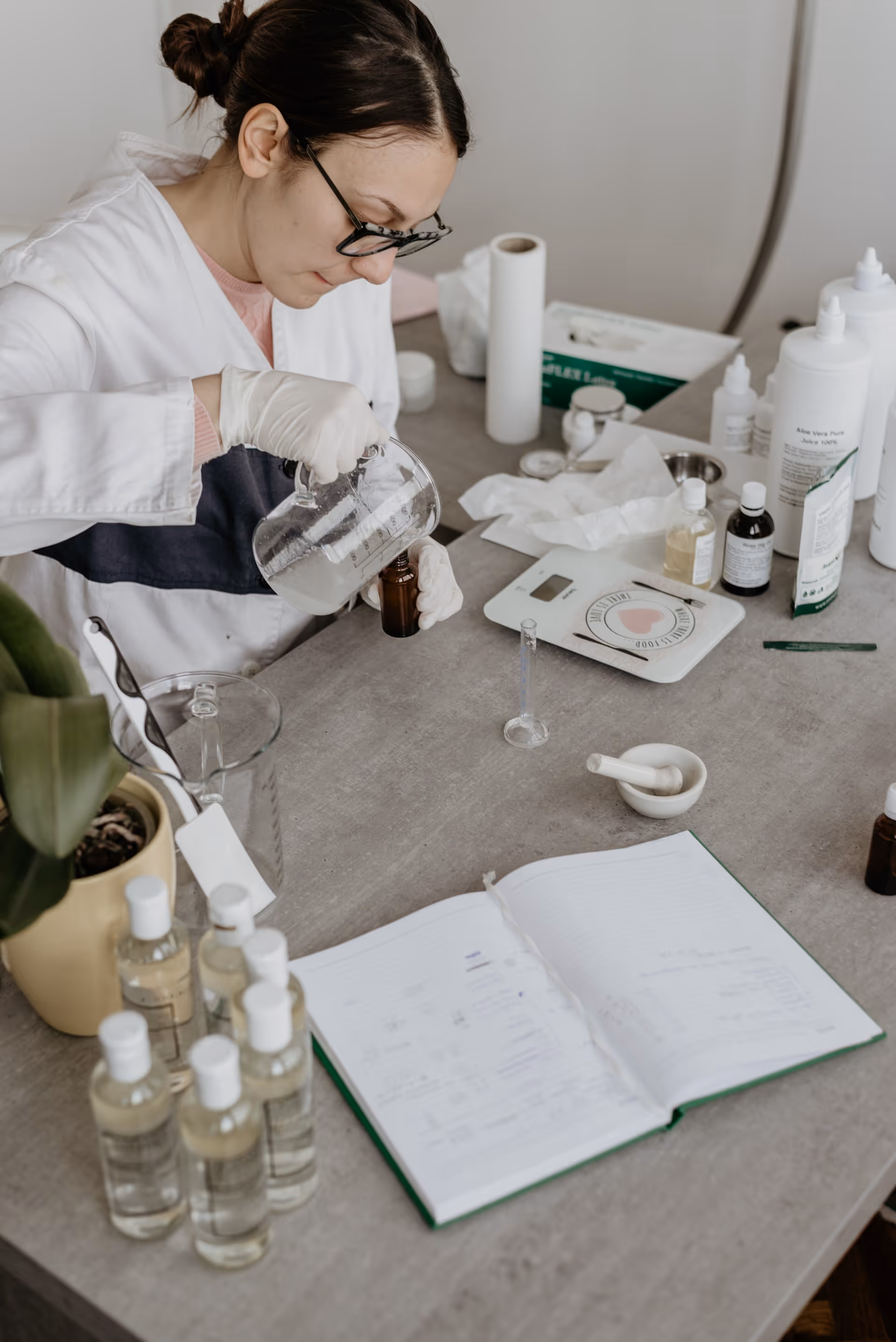 Woman in gloves mixing ingredients in a brown bottle on a lab table with bottles and a notebook.