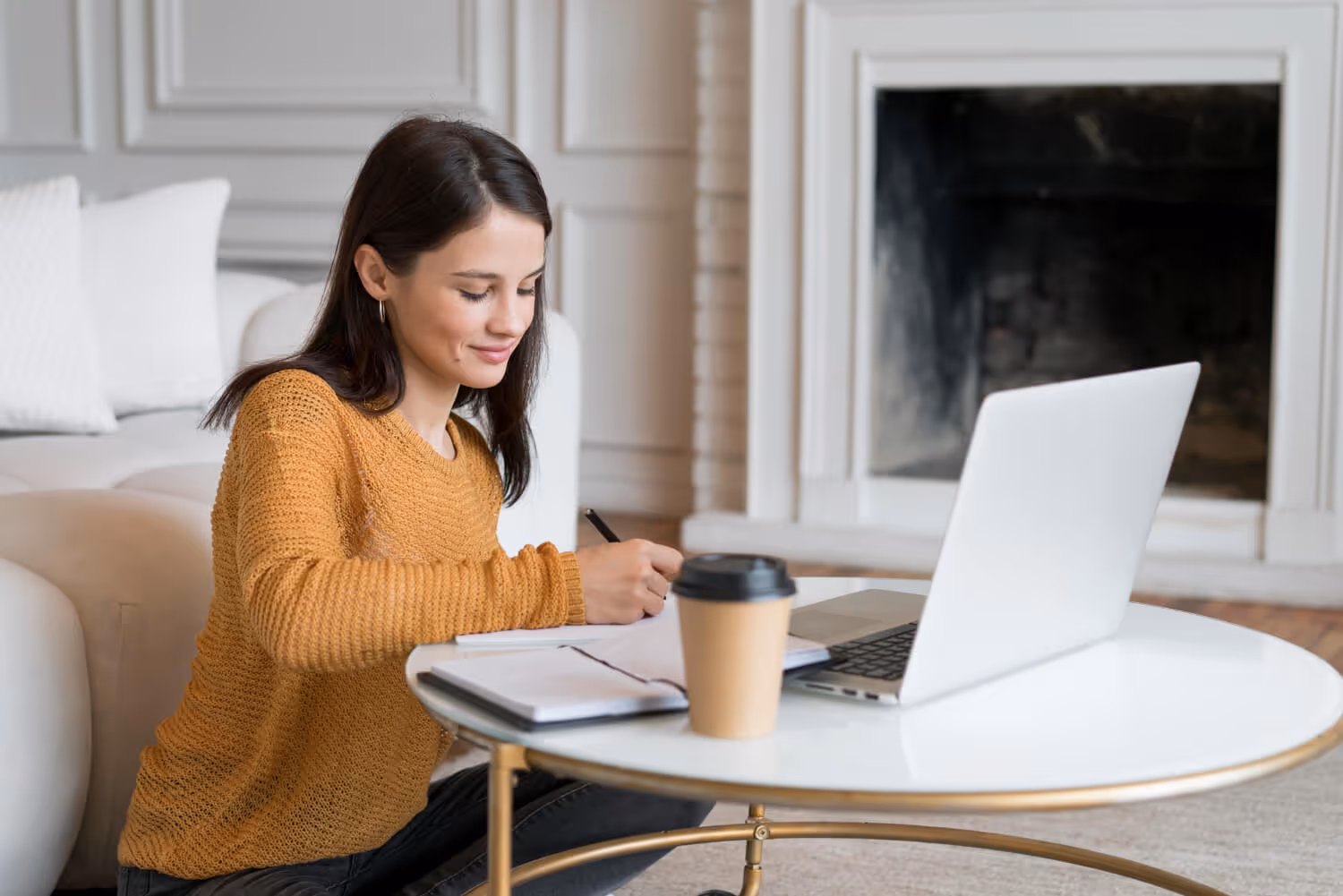 Woman in mustard sweater writing in notebook at round table with laptop and coffee cup nearby.