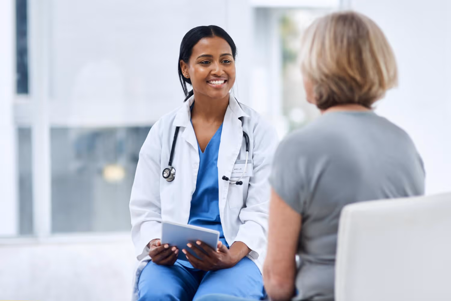Smiling female doctor in white coat and blue scrubs consulting with a seated patient in a bright clinic.