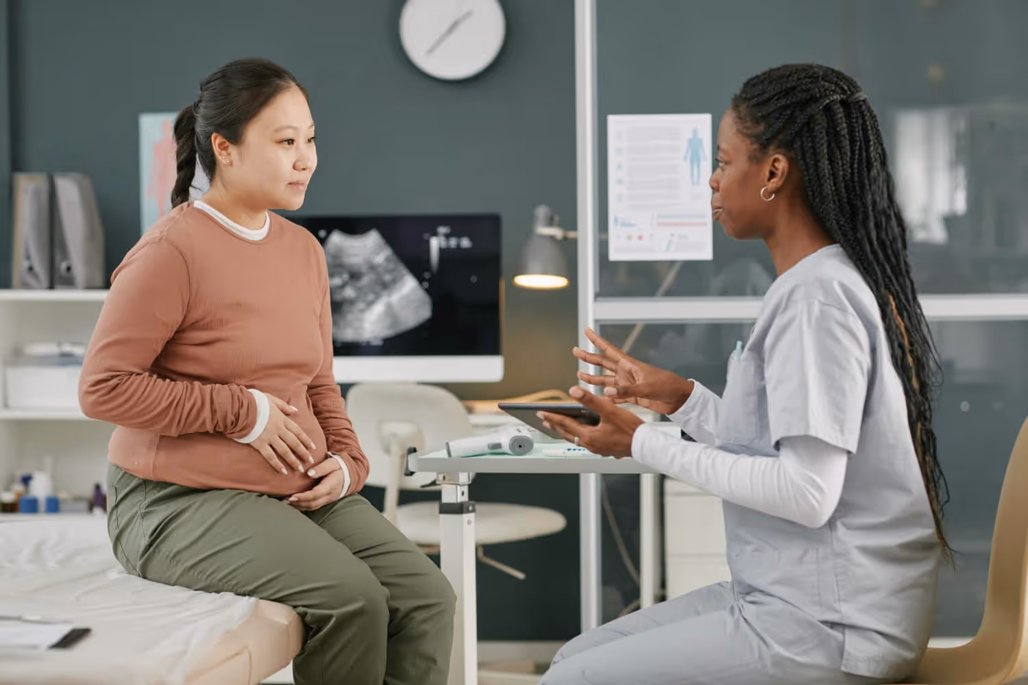 Pregnant woman sitting on exam table talking to healthcare professional holding a tablet in clinic.