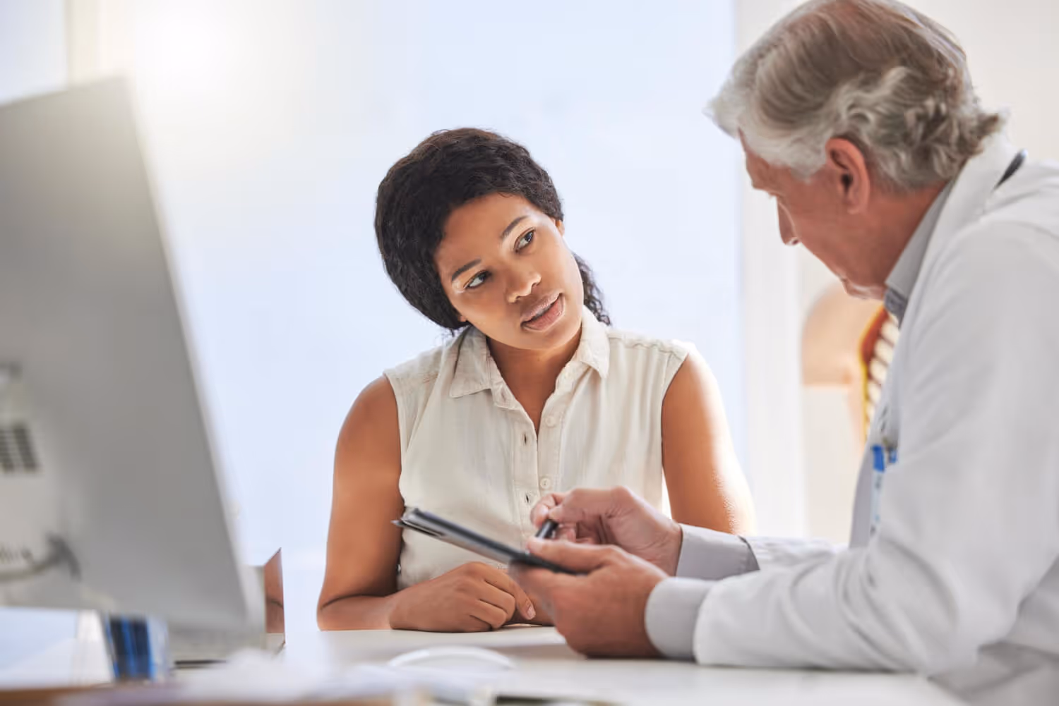 Woman attentively listening to an elderly doctor explaining notes on a tablet in a medical office.