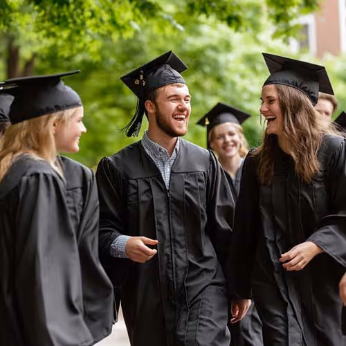 Group of graduates in caps and gowns smiling and talking outdoors with green trees in background.