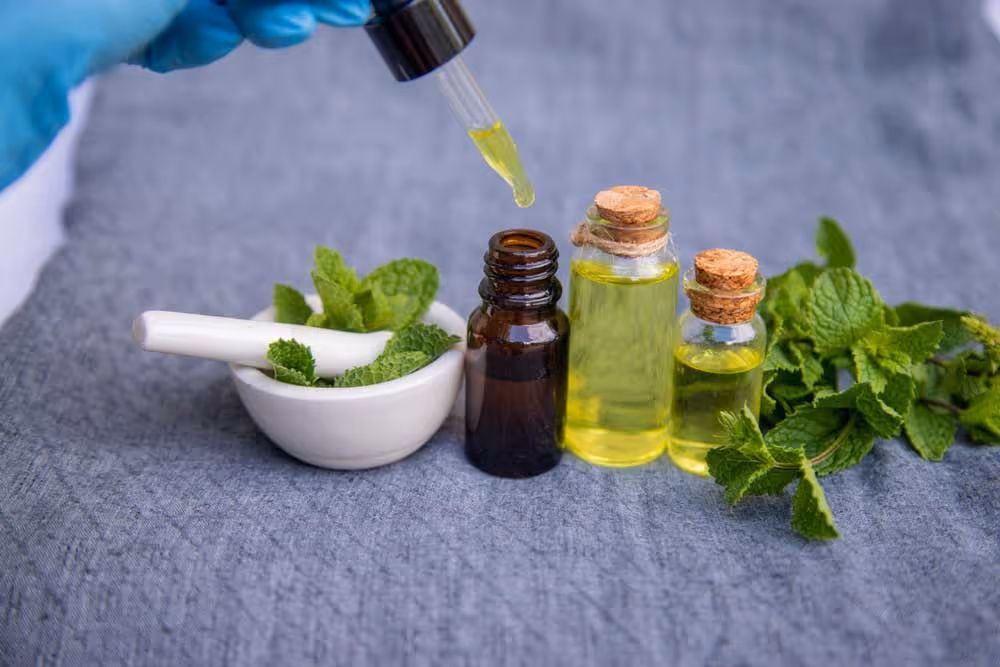 Dropper with yellow oil above small bottles and fresh mint leaves on gray fabric background.