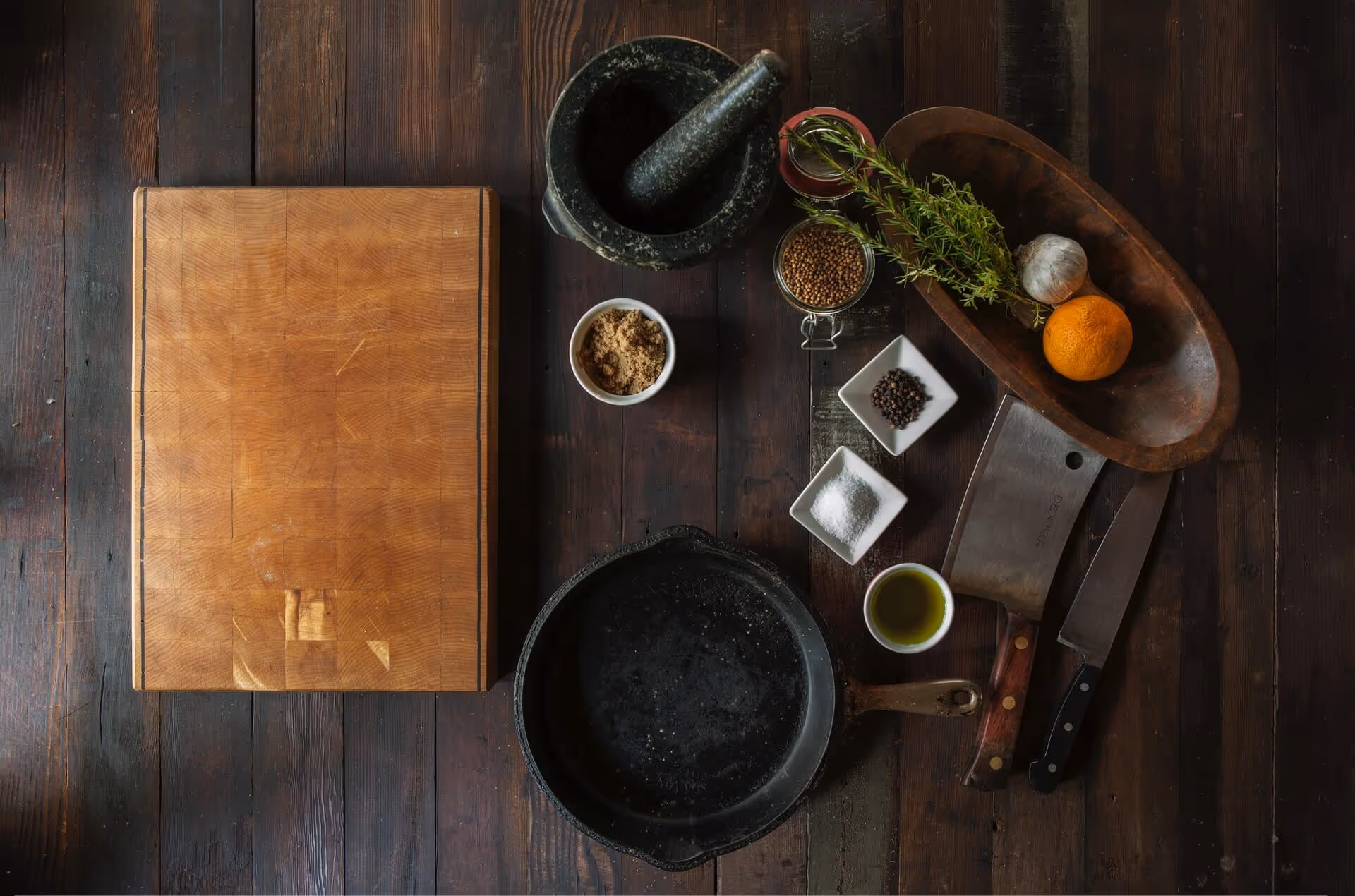 Wood cutting board, cast iron skillet, mortar and pestle, spices, herbs, knives, orange, and garlic on wood table.