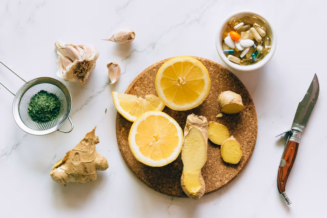 Lemon halves, ginger root slices, garlic cloves, dried herbs, pill capsules, and a knife on white surface.