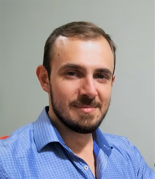 Man with short brown hair and beard wearing a blue patterned shirt against a plain wall.