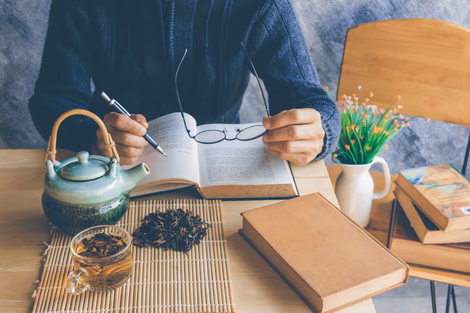 Person holding glasses and pen while reading an open book with tea and dried leaves on the table.