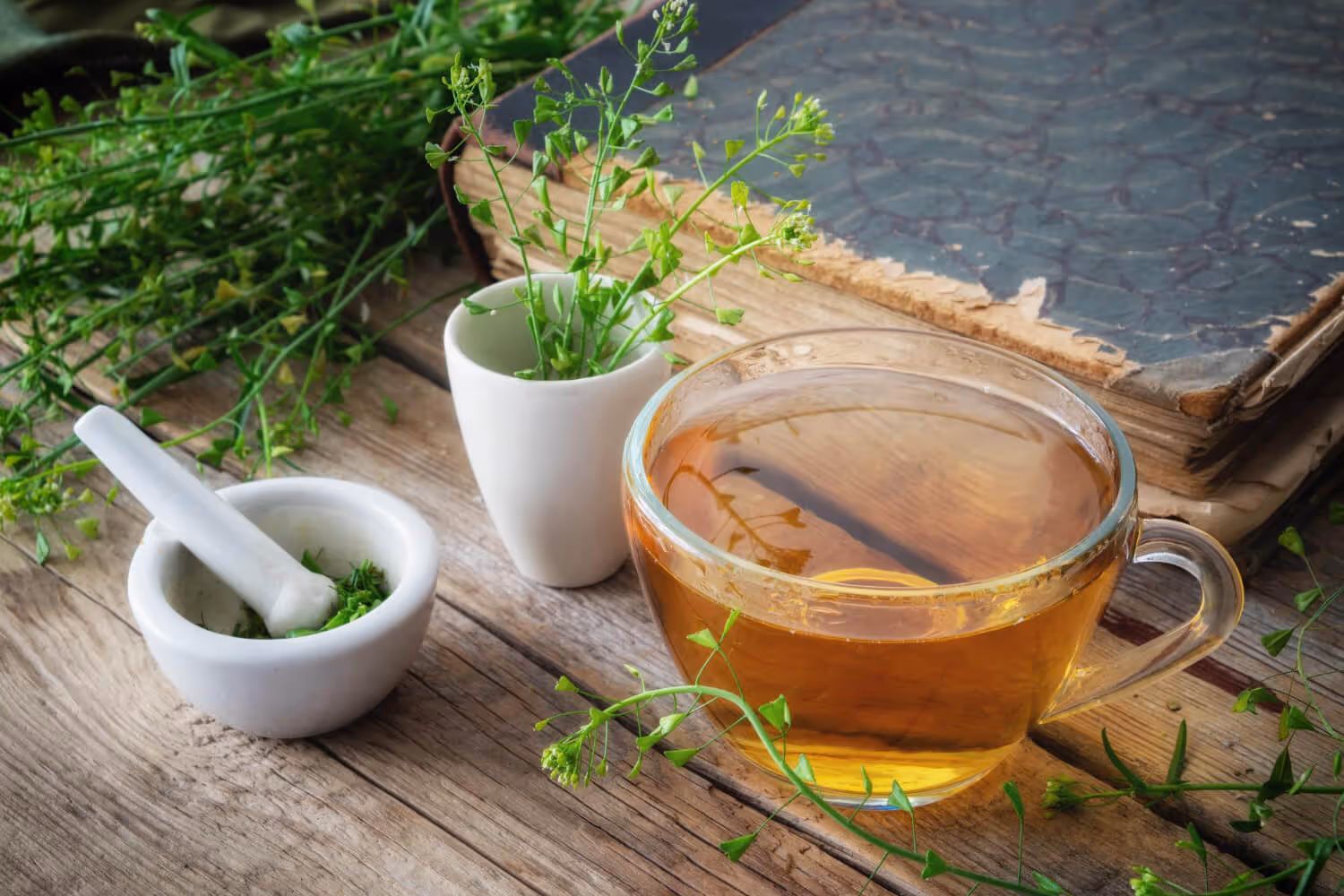 Glass cup of herbal tea with fresh green herbs, mortar and pestle, and an old book on a wooden table.