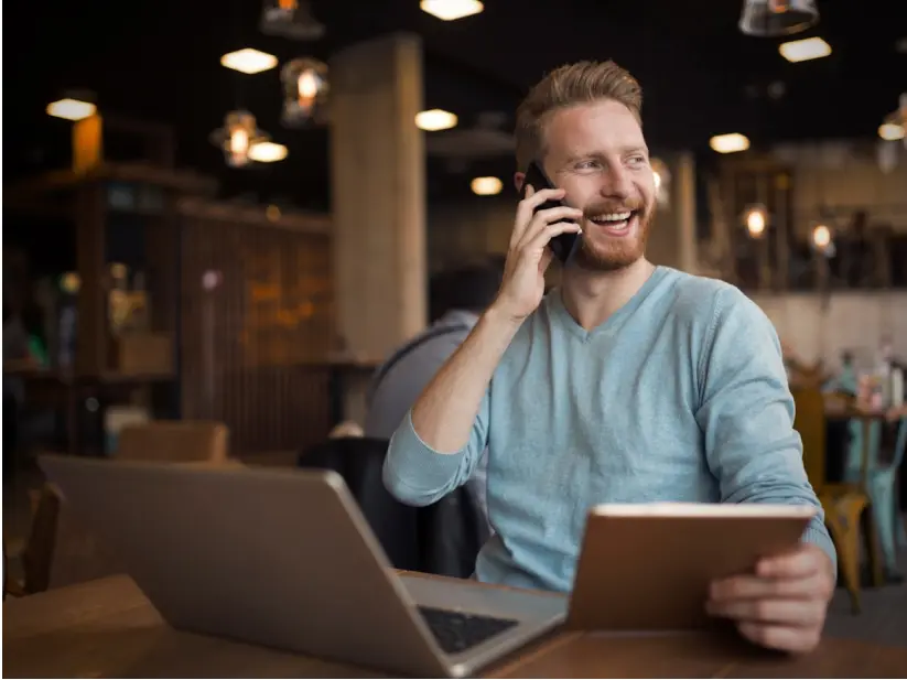 Man in blue sweater talking on phone while holding tablet at a table with laptop in a café.