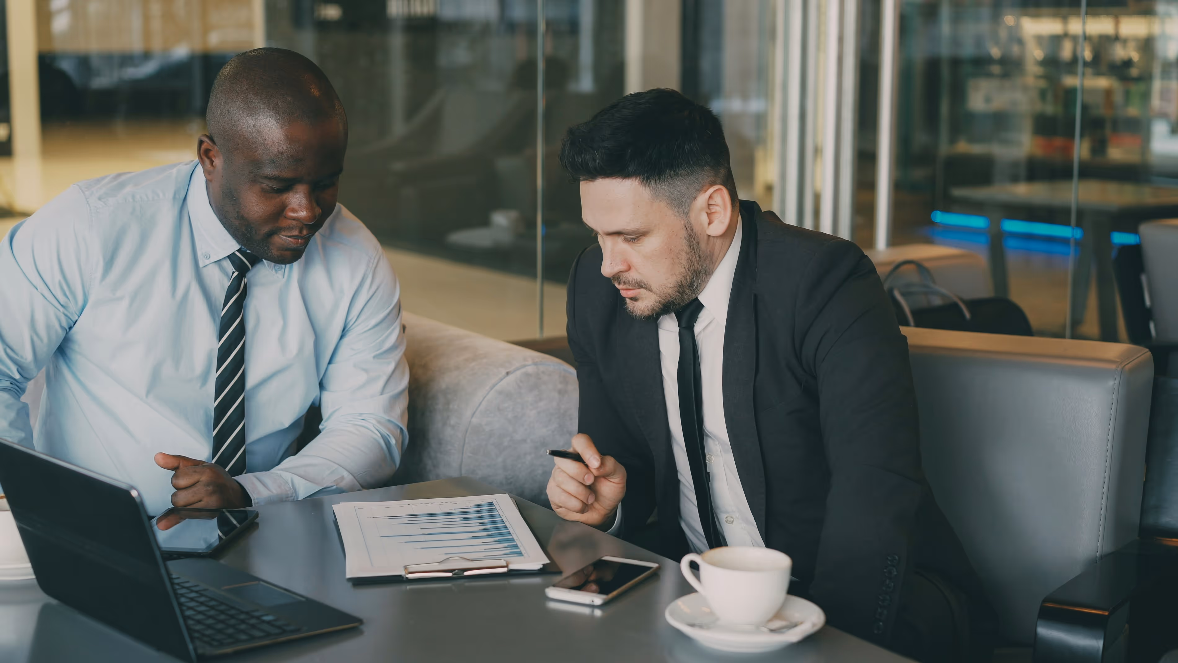 Two men in business attire discussing charts and data at a table with laptop, phone, and coffee cup.