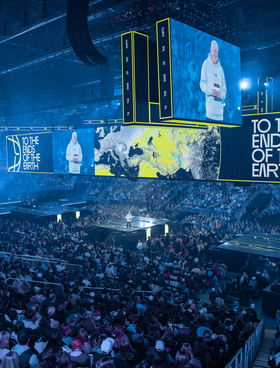 A large indoor arena filled with an audience watching a speaker on stage with multiple large screens displaying the event title 'To The Ends of the Earth' and a map of the Earth.