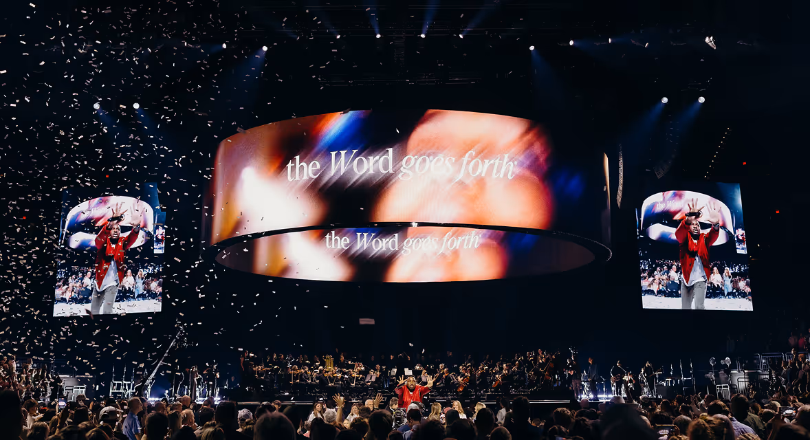 Crowd at concert with confetti falling and singer on stage in red jacket raising hands.