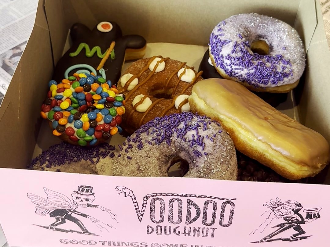 An open pink Voodoo Doughnut box with the Voodoo Doll, the M&M one, a maple bar and purple sprinkles