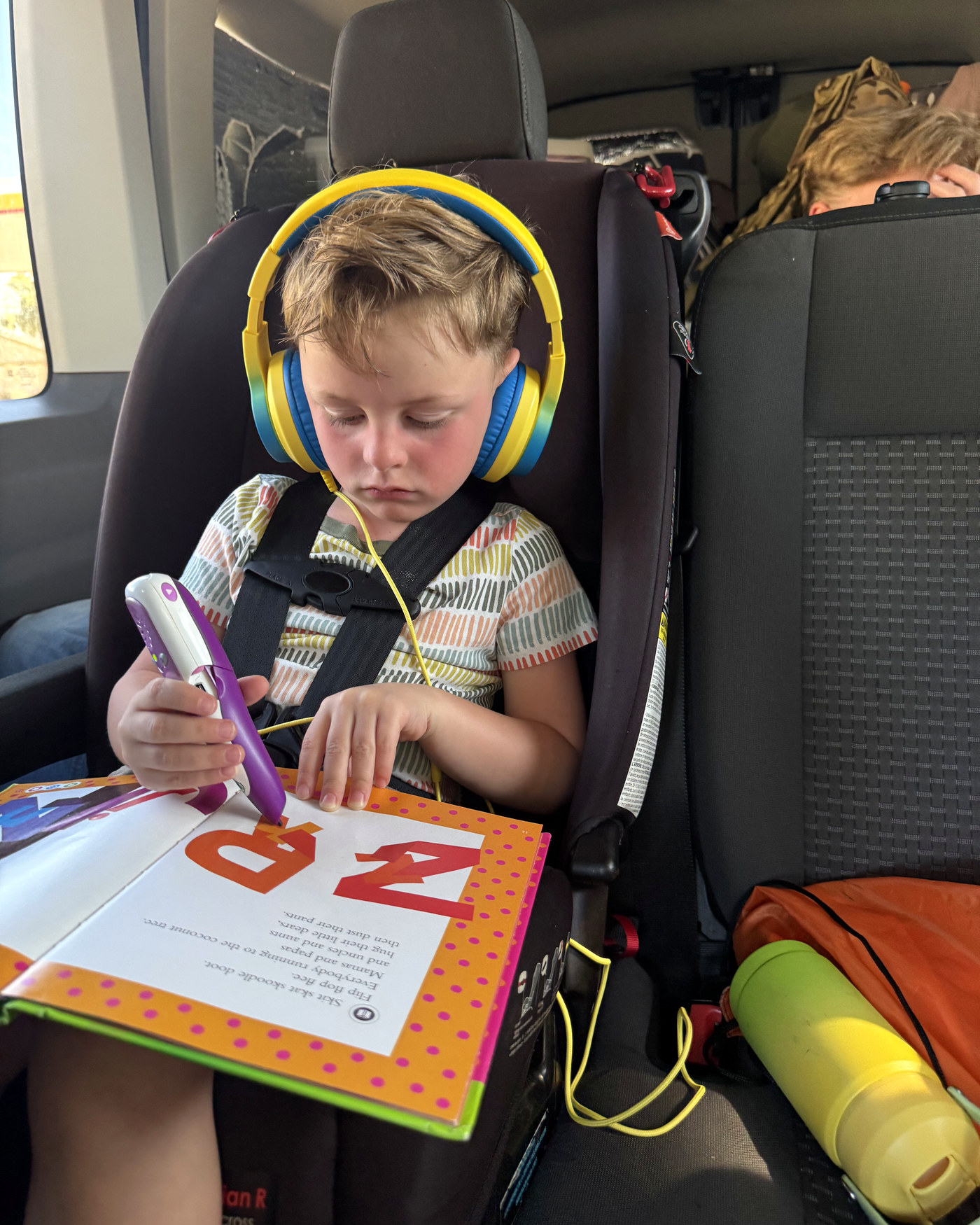 One of the boys in his car seat in the van, headphones on, reading a LeapFrog book with the pen