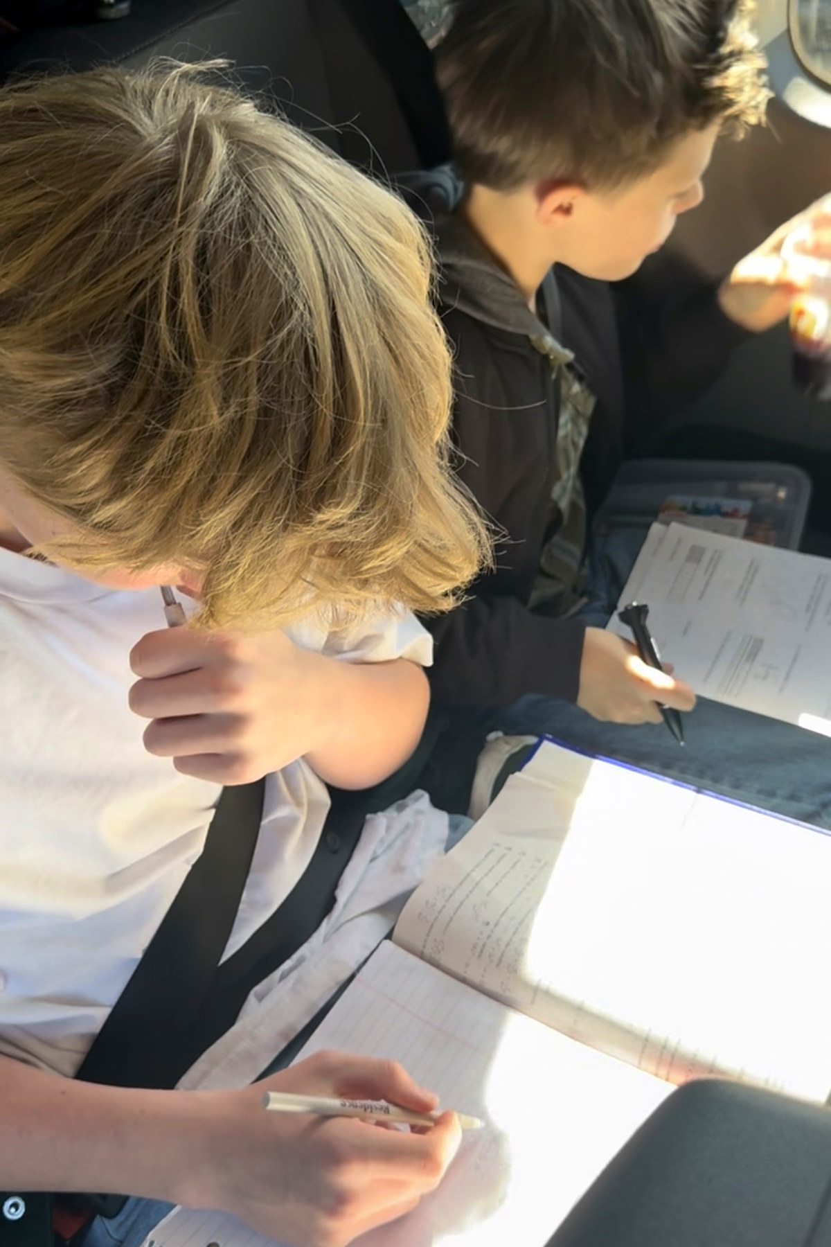 Two of the kids doing schoolwork in their seats in the van, notebooks open, sun coming through the window