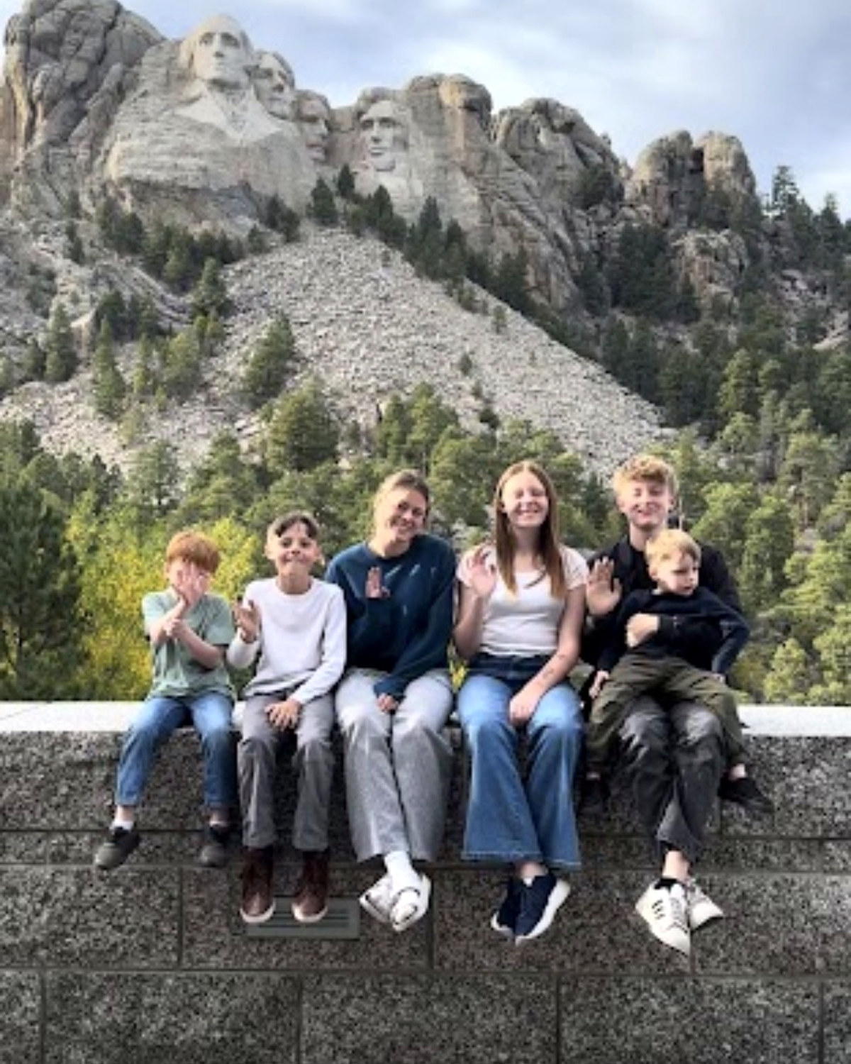 All six kids sitting on the wall at Mount Rushmore, waving, the presidents behind them