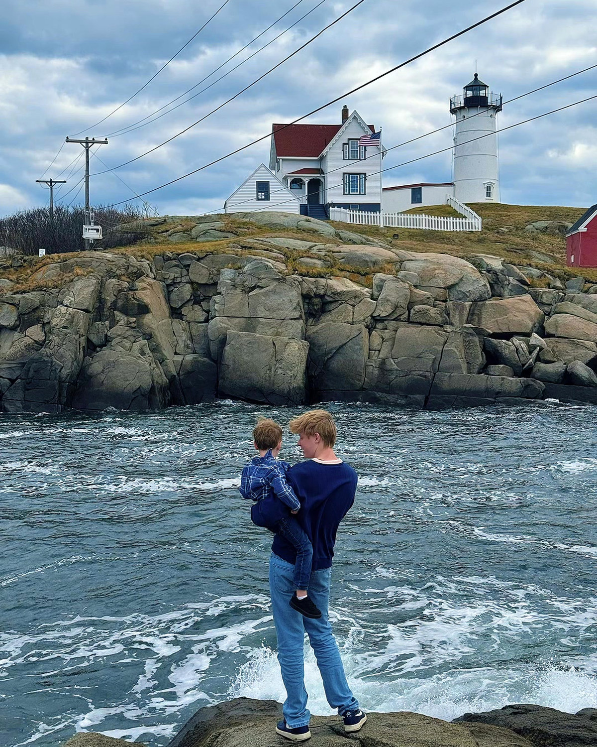 One of the boys holding his little brother on the rocks in York, Maine, the lighthouse behind them