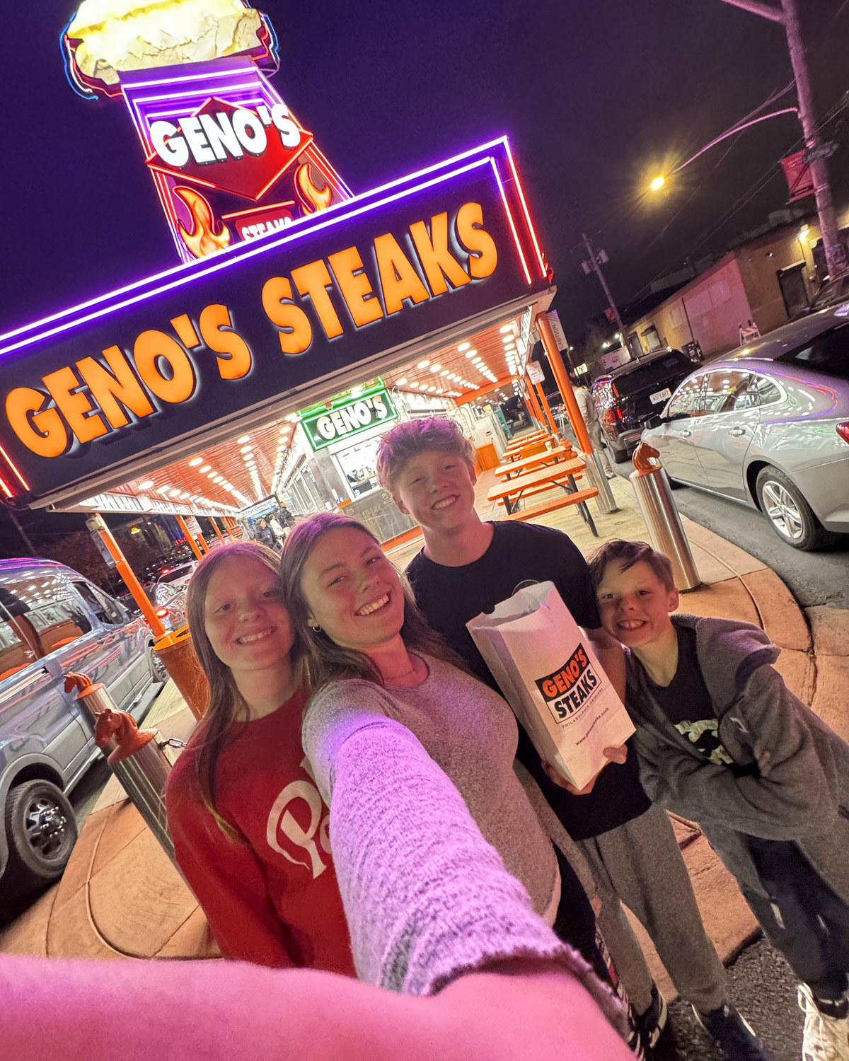 Four of the kids in a selfie under the neon Geno's Steaks sign at night