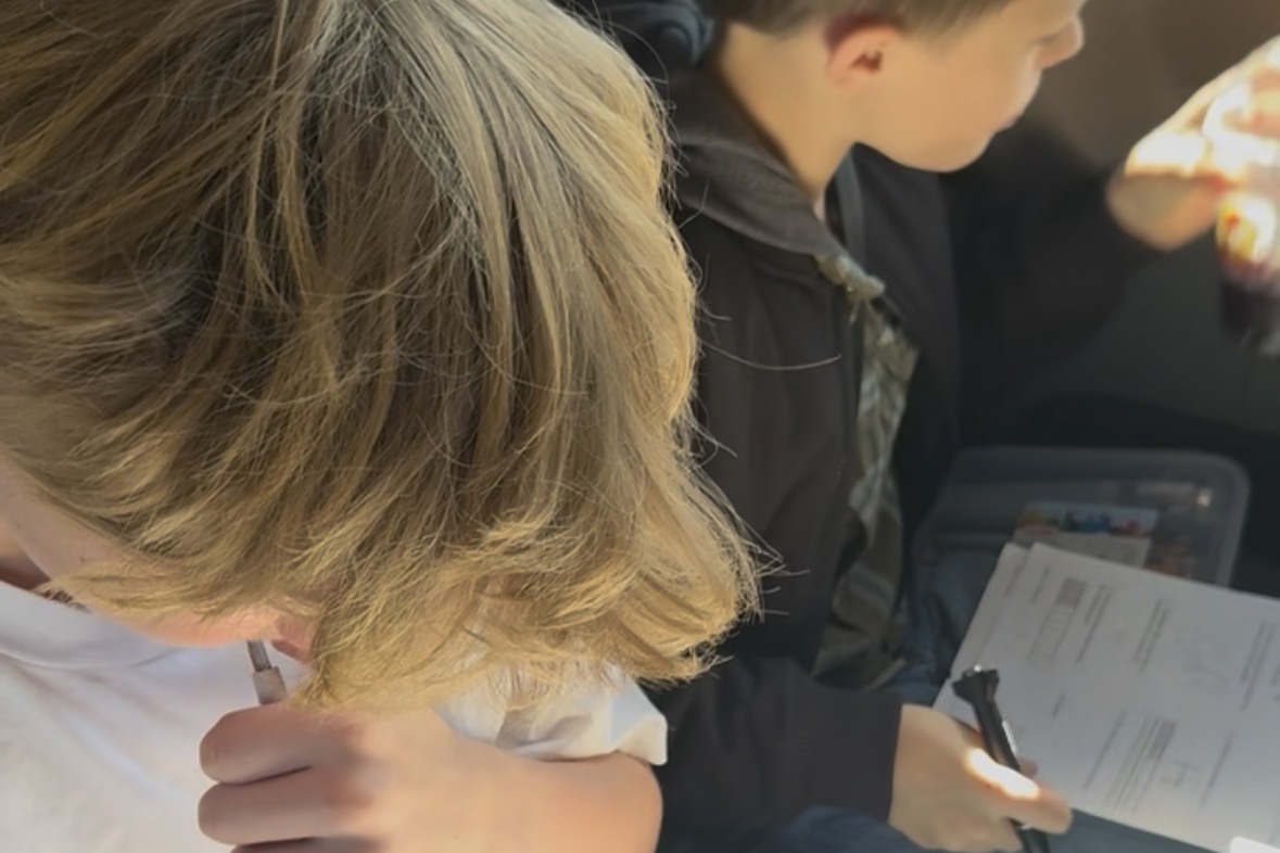 Two of the kids doing school in their van seats on a road trip, notebooks on their laps and sun coming in the window