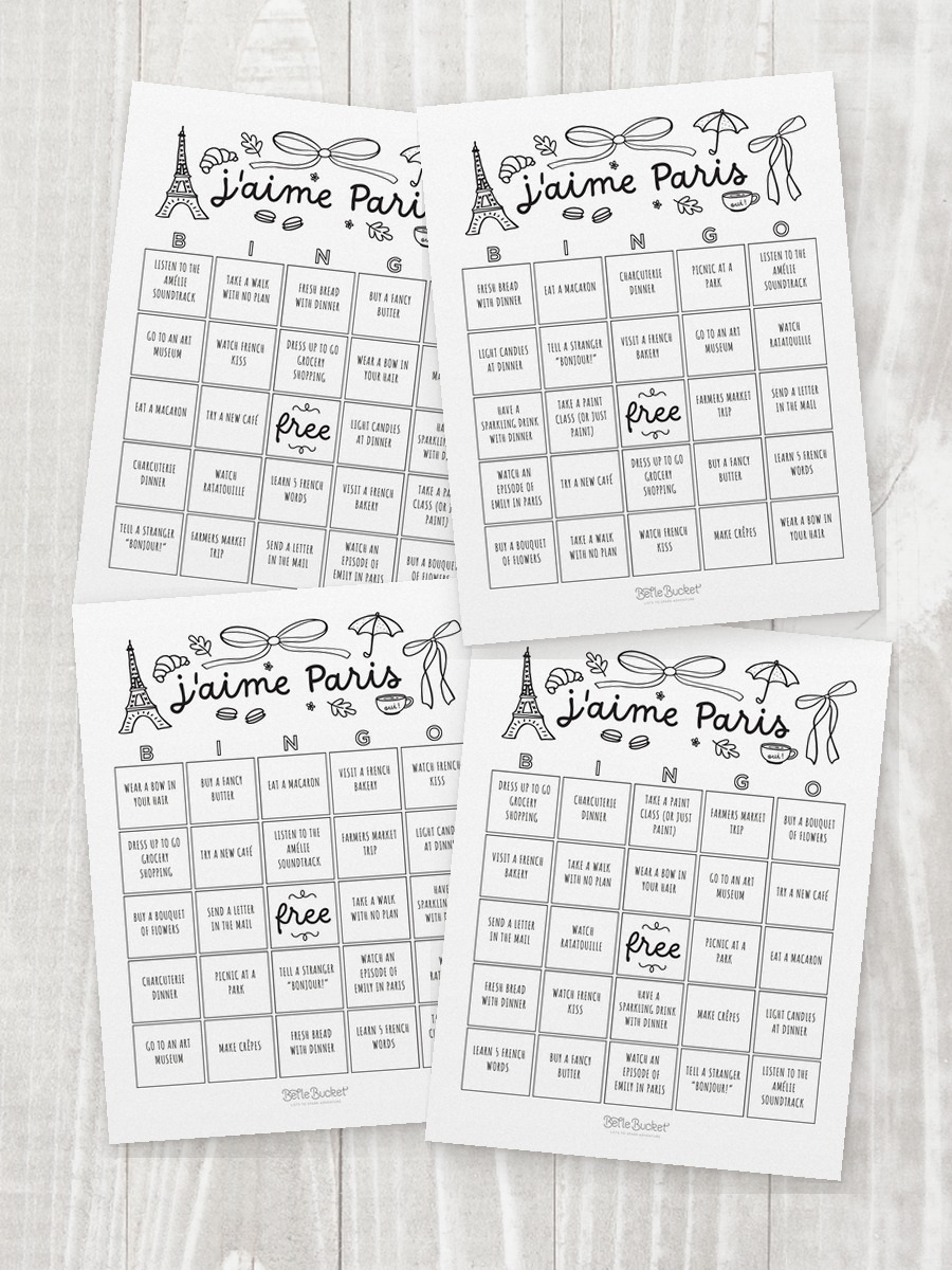 Four shuffled j'aime Paris bingo cards overlapping on a table, each with the squares in a different order