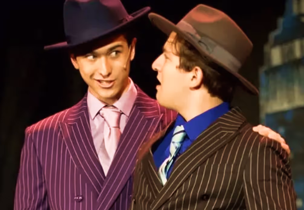 Two men wearing pinstripe suits and fedora hats, one with a purple suit and pink tie, the other with a brown suit and striped tie, engaged in conversation.