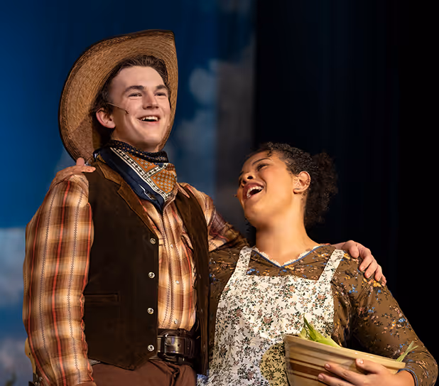 Two actors in vintage Western costumes smiling and embracing on stage, one holding a basket with vegetables.