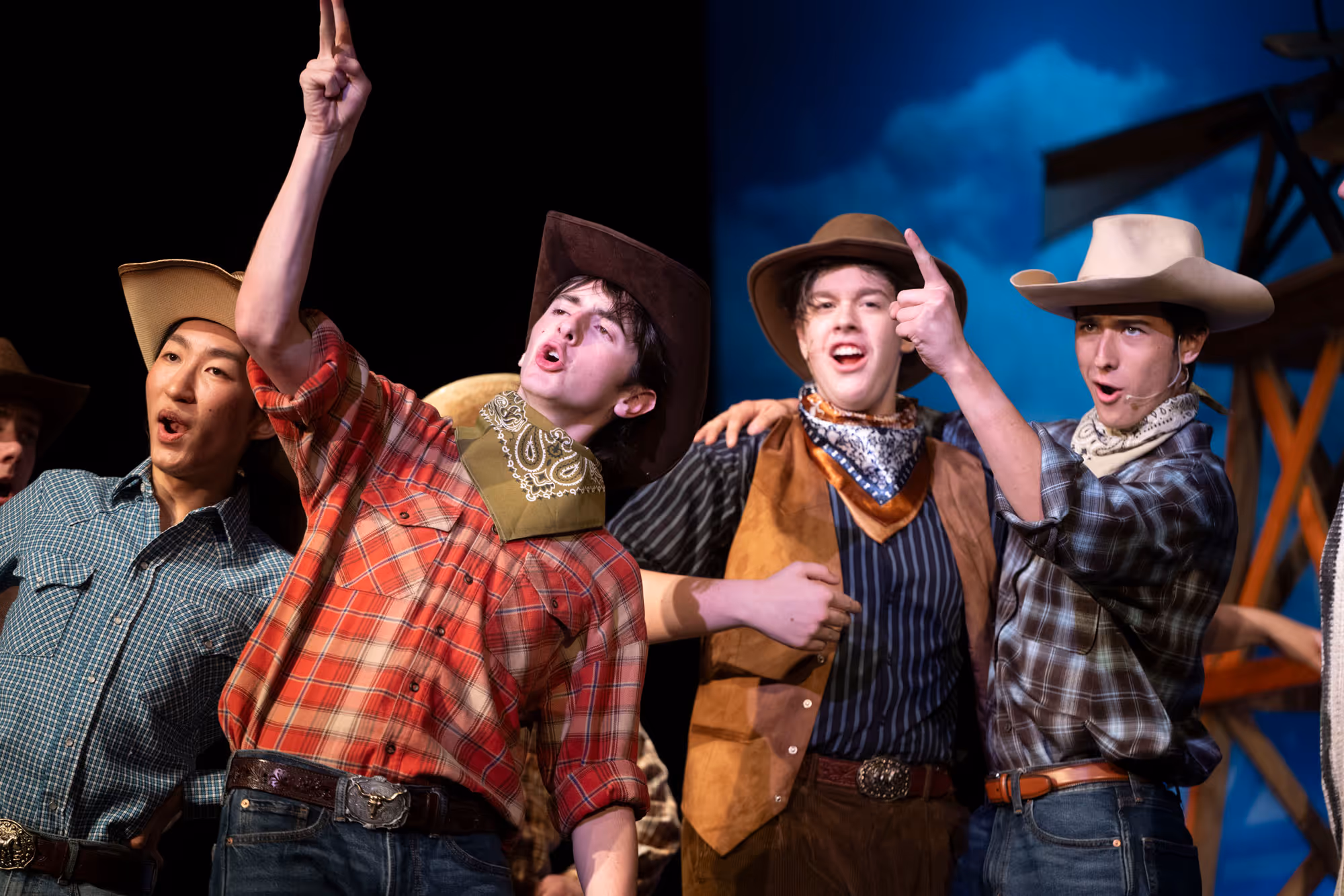 Actors in Western-style costumes performing on stage with a windmill backdrop under a blue sky.