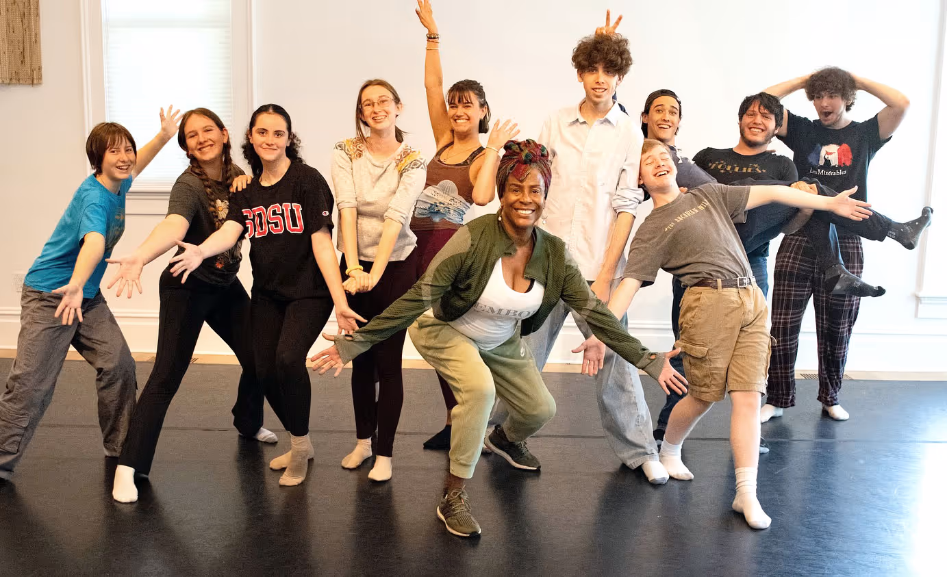 Group of smiling diverse young people and an adult posing playfully in a dance studio with open arms.