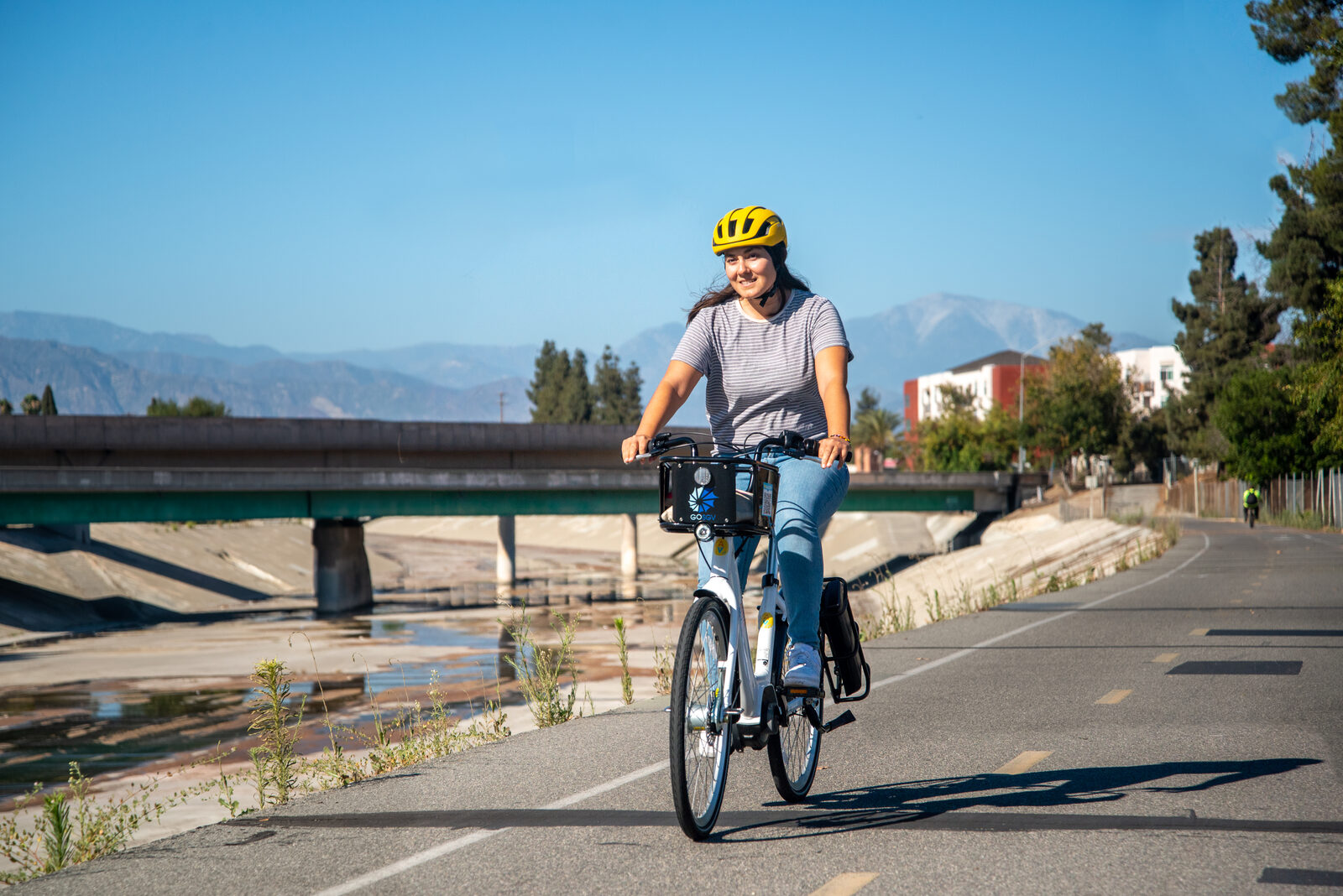 A woman rides a white GoSGV commuter e-bike on a paved river path with mountains behind her.