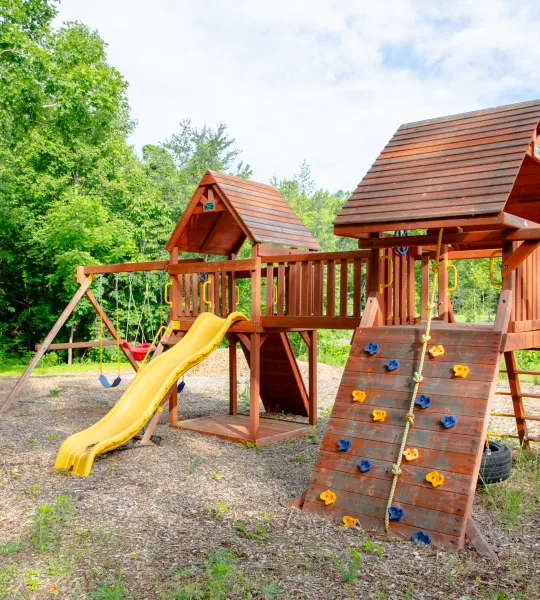 A wooden playground set with a slide, climbing wall, and swings in a sunny outdoor setting.