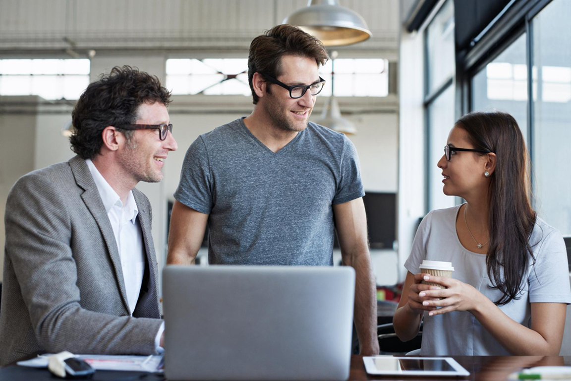 Three colleagues in casual office setting having a conversation around a laptop, one holding a coffee cup.