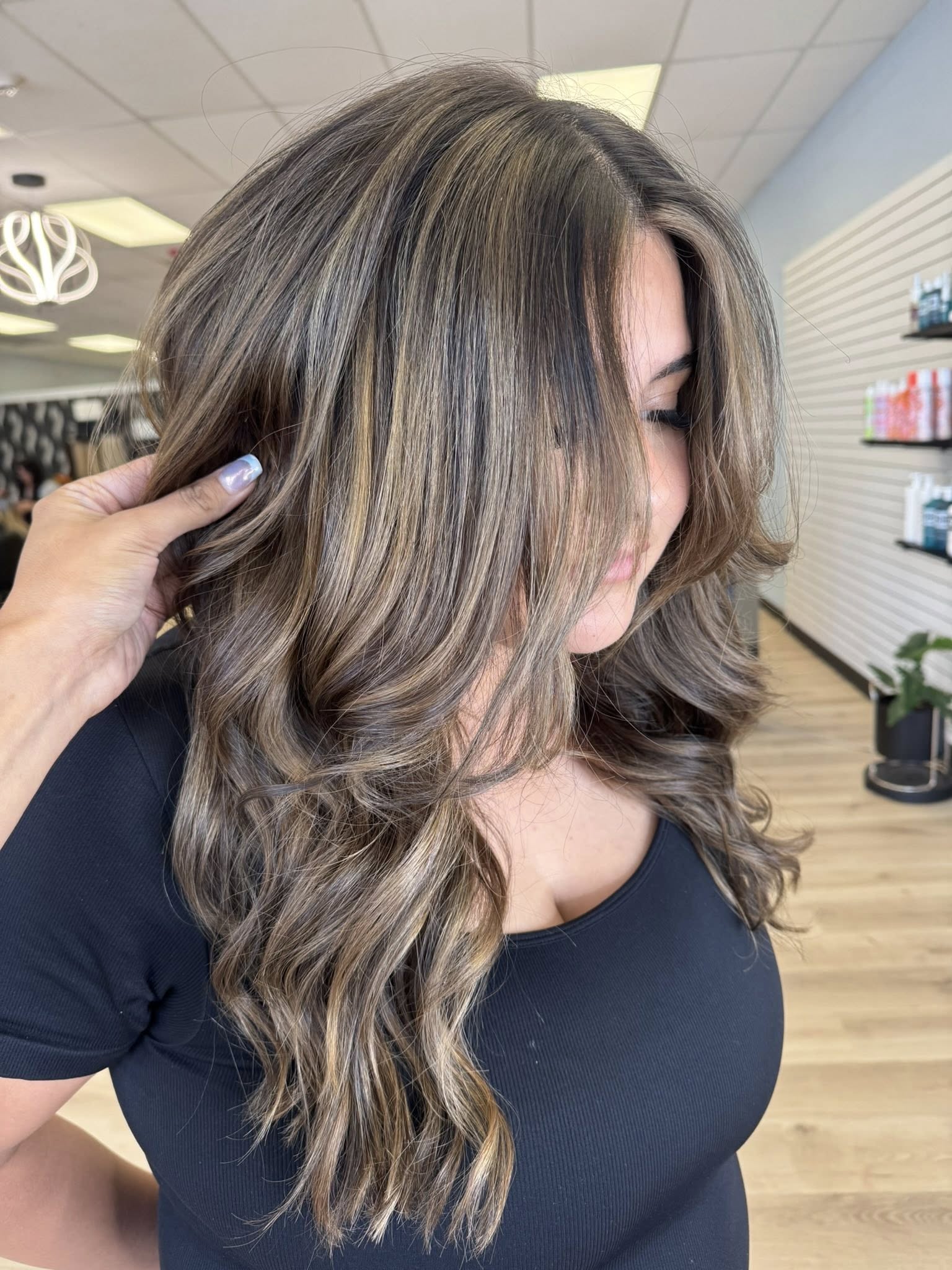 Woman with long, wavy hair styled with soft brown and blonde highlights inside a hair salon.