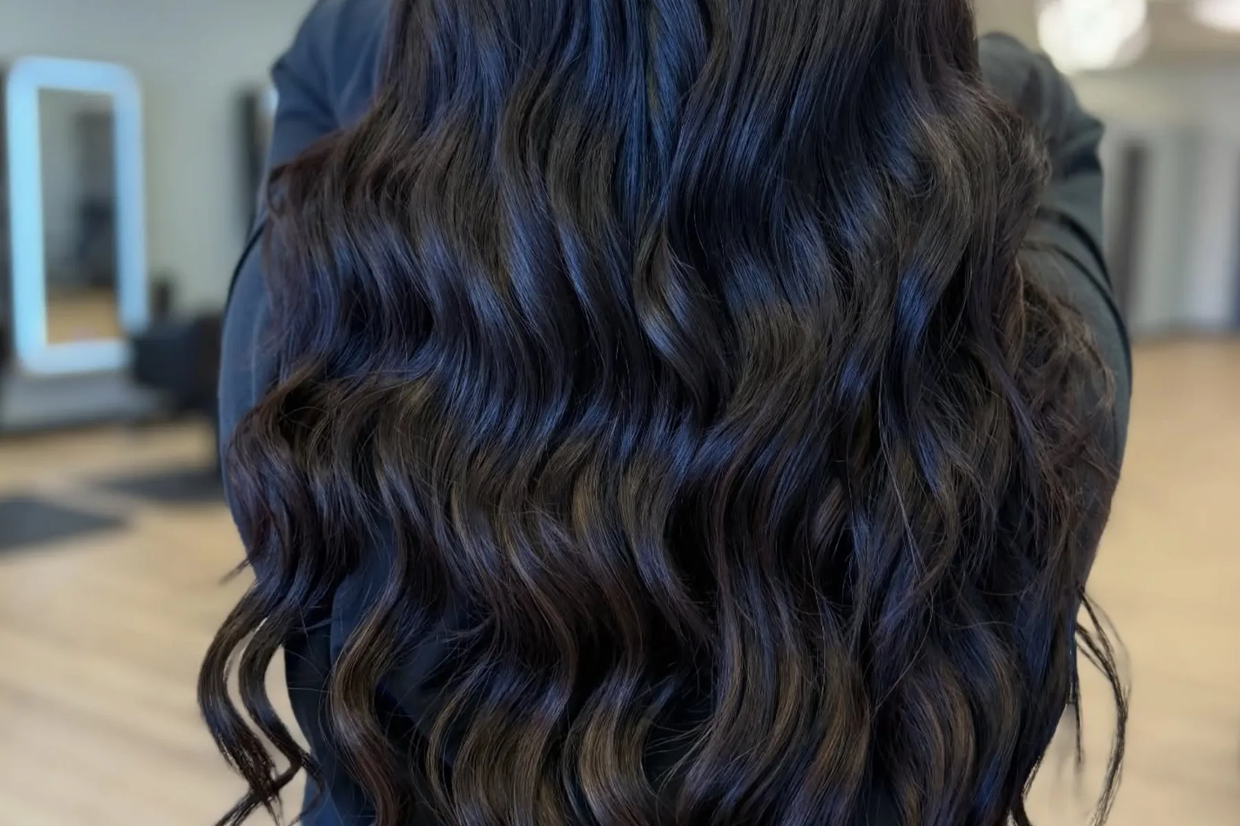 Long, wavy black hair with a healthy shine, viewed from the back in a salon setting.