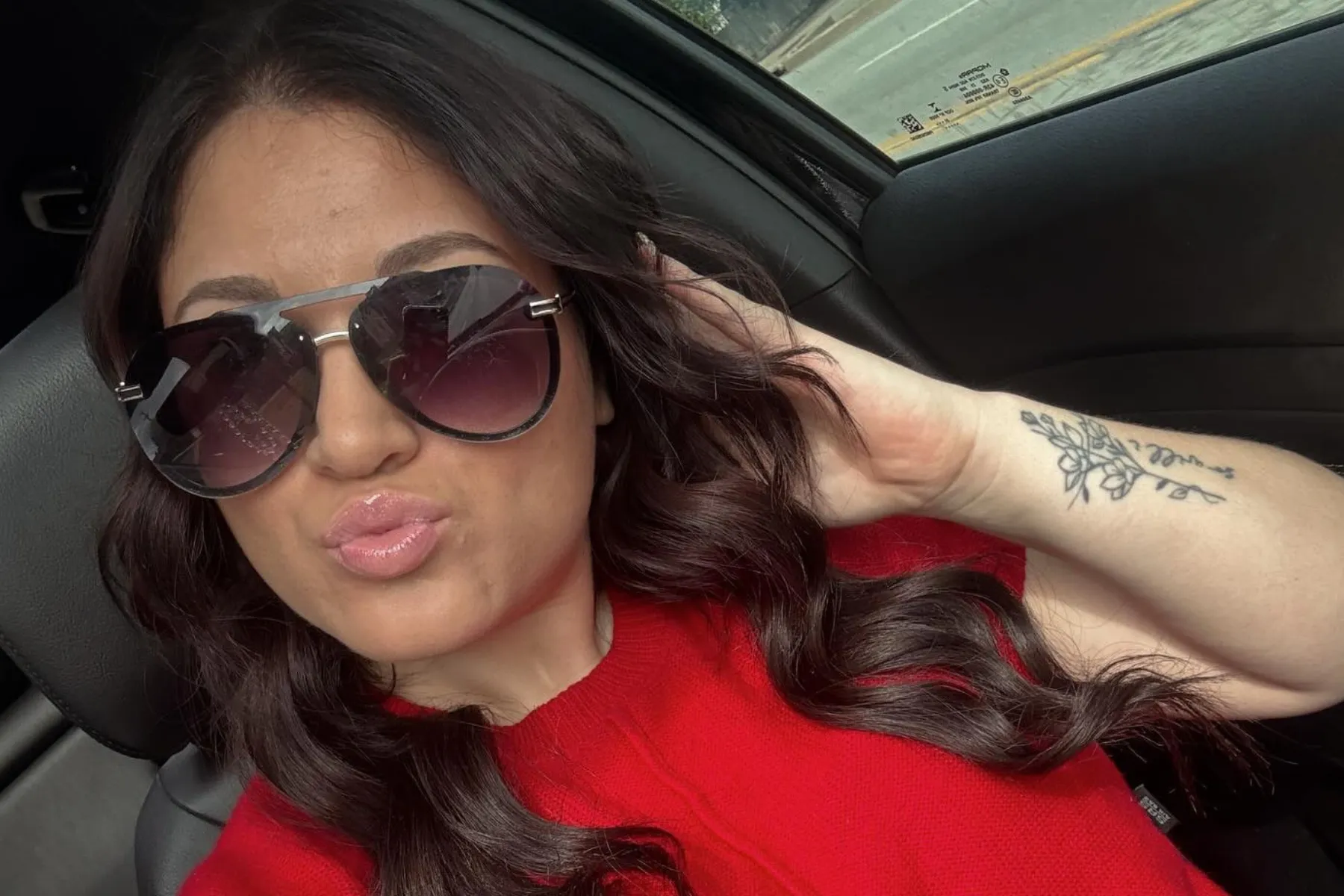 Woman with long dark wavy hair wearing a red top and large dark sunglasses, posing inside a car with a floral tattoo on her forearm.