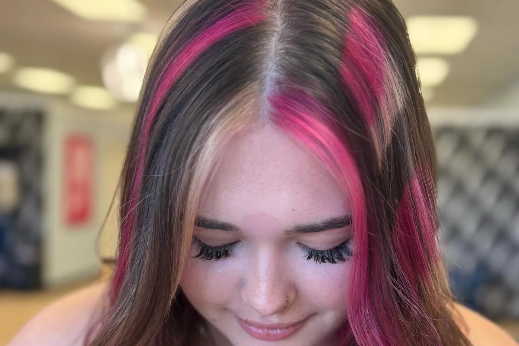 Woman with dark brown hair and vibrant pink highlights looking down with eyes closed indoors.